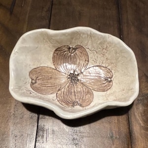 May include: A small, irregularly shaped ceramic dish with a cream-colored, textured surface. The center of the dish features a detailed, brown, four-petaled flower design with visible veining and a dark, textured stamen in the middle.