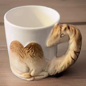 May include: A white ceramic mug featuring a three-dimensional camel relief on the side. The camel's body forms the base of the handle, while its long neck and head curve upward to create the handle itself, complete with painted closed eyes.