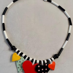 May include: A necklace featuring a strand of alternating black and white cylindrical beads. The center of the necklace is adorned with five heart-shaped charms in yellow, light green with black crosses, solid red, black with pink dots, and orange.