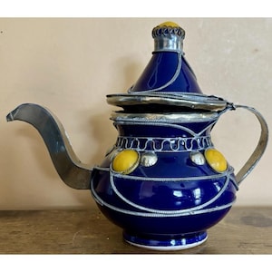 May include: A deep blue teapot featuring a conical lid with a yellow knob. The body is adorned with silver-toned metal wire detailing, a curved spout, a handle, and two prominent yellow oval accents set within the metallic framework.