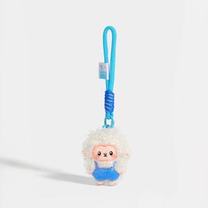 May include: A plush sheep keyring featuring a white curly fleece texture, a pink face with black eyes, and blue dungarees. The figure is attached to a bright blue woven cord strap with a matching blue knot detail and a small white fabric tag.