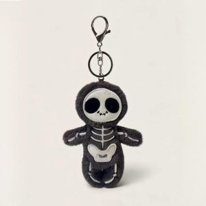 May include: A plush skeleton keychain featuring a dark gray fuzzy body with white embroidered bone details. The character has a round head with large black circular eyes and a small stitched mouth. It is attached to a silver metal lobster clasp.
