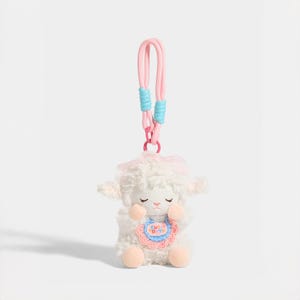 May include: A plush white lamb keyring featuring a pink bow on its head and a crocheted bib with the text "CUT DAYS" in colourful letters. The lamb has closed eyes, peach-coloured paws, and is attached to a pink cord with light blue accents.
