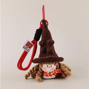 Op de afbeelding: Een gehaakte sleutelhanger met een bruine puntmuts met een textuur, een klein gezichtje met roze wangen en een rood-geel patroon sjaaltje. Het figuurtje zit vast aan een rood koord met een zwart accent en een rode metalen karabijnhaak.
