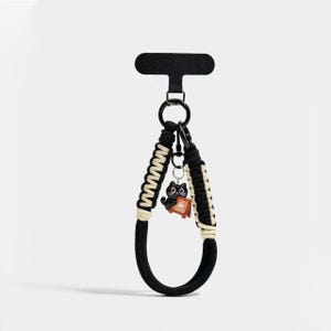 以下が含まれることがあります： 黒とクリーム色のマクラメ編みのリストストラップキーホルダー。黒い金属製のリングと、Coffeeと書かれた茶色の看板を持つ黒猫の小さなチャームが付いています。ストラップは黒いT字型の台紙に取り付けられています。