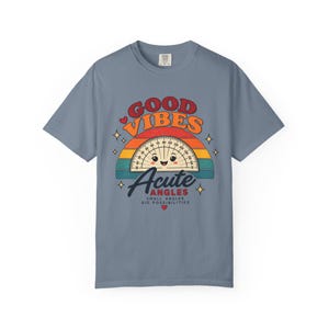 May include: A slate blue short-sleeve t-shirt featuring a graphic of a smiling protractor under a rainbow arch. The text reads GOOD VIBES, ACUTE ANGLES, and SMALL ANGLES BIG POSSIBILITIES, accented with small stars and red hearts on the front.