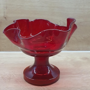 Könnte beinhalten: Eine leuchtend rote Glasschale mit einem breiten, gerüschten und gewellten Rand. Die Schale wird von einem stabilen, kreisförmigen Sockel getragen, was eine klassische Fußform ergibt. Das durchscheinende rote Glas hat eine glatte Oberfläche.