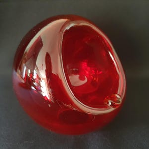 Pode incluir: Um objeto esférico de vidro vermelho com uma superfície lisa e polida e uma grande abertura circular rebaixada em um lado. O vidro é translúcido, permitindo a passagem de luz, e apresenta uma pequena conta de vidro transparente na borda.