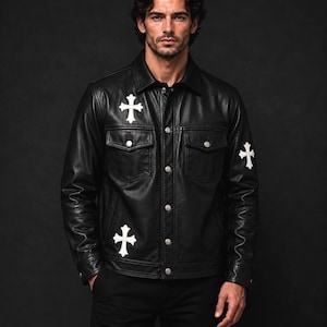 May include: A black leather jacket featuring a pointed collar, long sleeves with buttoned cuffs, and a front button closure. The jacket is adorned with white cross-shaped patches on the chest, lower front, and sleeve, and includes two chest pockets.