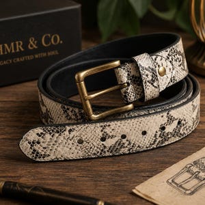 May include: A belt featuring a cream and black snakeskin pattern with a brass-toned metal buckle. The belt is coiled and includes a black interior lining. A black box with the text AHMR & CO. A LEGACY CRAFTED WITH SOUL is visible nearby.