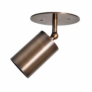 May include: A bronze-toned cylindrical spotlight fixture featuring a circular mounting plate with two visible screw holes. The light head is connected to the base by a swivel joint, allowing for adjustable positioning of the light source.