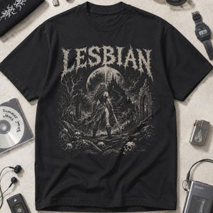 Può includere: Una t-shirt nera a maniche corte con una stampa grafica bianca e la scritta LESBIAN in alto. L'illustrazione raffigura una figura che impugna una spada davanti a un castello e una grande luna, circondata da alberi, rovine e teschi.