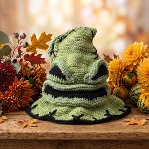 May include: A light green crocheted hat featuring a pointed top, a wide brim, and black yarn detailing that mimics facial features including eyes and a mouth. The hat has visible black cross-stitch patterns along the brim and upper section.