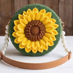 Puede incluir: Un bolso redondo de ganchillo verde oscuro con una gran aplicación de girasol tridimensional con centro marrón y pétalos amarillos brillantes. El bolso incluye una correa hecha de perlas blancas artificiales y un material liso de color canela.