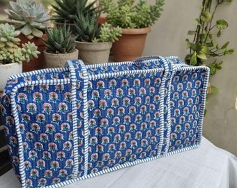 May include: A rectangular quilted tote bag in blue featuring a repeating pattern of small pink and green floral motifs. The bag has two long handles and white and blue striped fabric trim along the edges, seams, and the top opening.