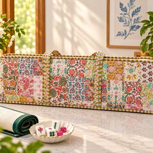May include: A rectangular, quilted fabric carrying case featuring a vibrant patchwork of floral patterns in pink, orange, green, and blue. The case has two handles and green and white striped trim. Nearby sits a small bowl with mahjong tiles and dice.