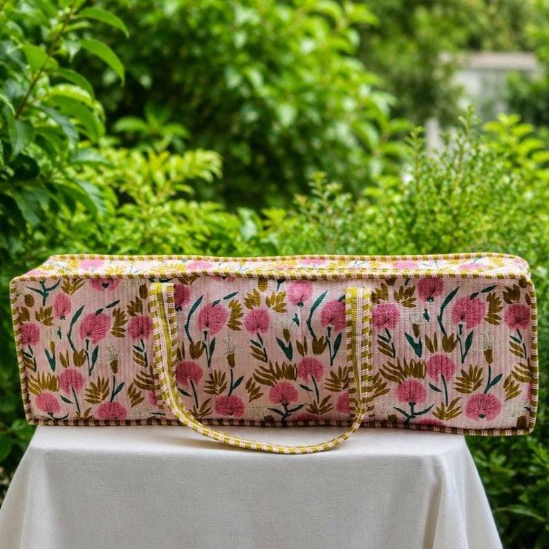 May include: A rectangular fabric bag featuring a light pink base with a repeating pattern of dark pink flowers and green leaves. The bag has a structured shape with a flat top and bottom, finished with a green and white striped fabric trim.