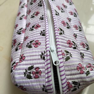 May include: A quilted fabric pouch featuring a pattern of small pink flowers with green leaves on a background of thin white and light purple horizontal stripes. The pouch includes a white zipper closure with a YKK branded metal pull tab.