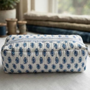 May include: A rectangular quilted zippered pouch featuring a white base with a repeating blue paisley print. The top closure includes a light blue and white striped fabric border along the zipper line, providing a structured shape.