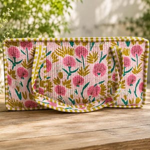 May include: A rectangular quilted tote bag featuring a light pink base with a repeating floral pattern in shades of dark pink, olive green, and teal. The bag has two matching handles and is finished with a striped olive green and white trim.