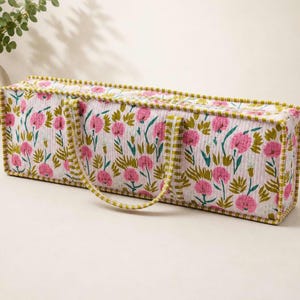 May include: A rectangular quilted storage bag featuring a floral pattern with pink flowers and green leaves on a white background. The bag has two fabric handles and is finished with a green and white striped trim along all the edges.