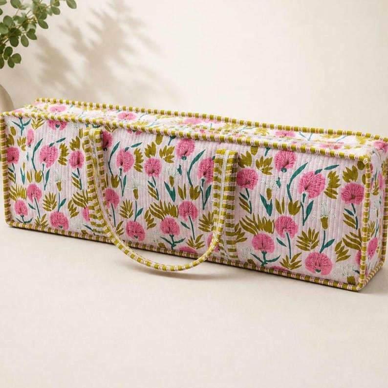 May include: A rectangular quilted storage bag featuring a floral pattern with pink flowers and green leaves on a white background. The bag has two fabric handles and is finished with a green and white striped trim along all the edges.