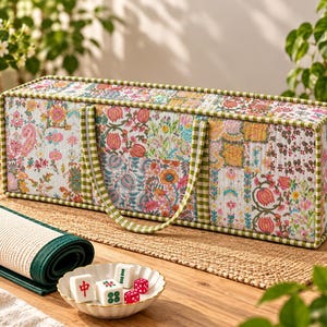May include: A rectangular quilted fabric storage case featuring a colorful floral pattern with green and white gingham trim and two matching handles. Beside it sits a rolled green and white mat and a small white bowl containing mahjong tiles.