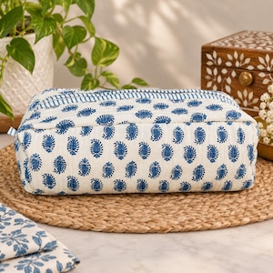May include: A rectangular quilted pouch featuring a white base with a repeating blue paisley print and a blue striped border along the top edge. The fabric has a soft, textured appearance with visible stitching lines throughout the entire surface.
