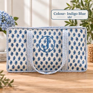 May include: A rectangular quilted tote bag in off-white featuring a repeating indigo blue paisley pattern. The bag has blue and white striped trim and handles, with a large embroidered indigo blue letter J inside a circle. Text reads Colour- Indigo Blue.