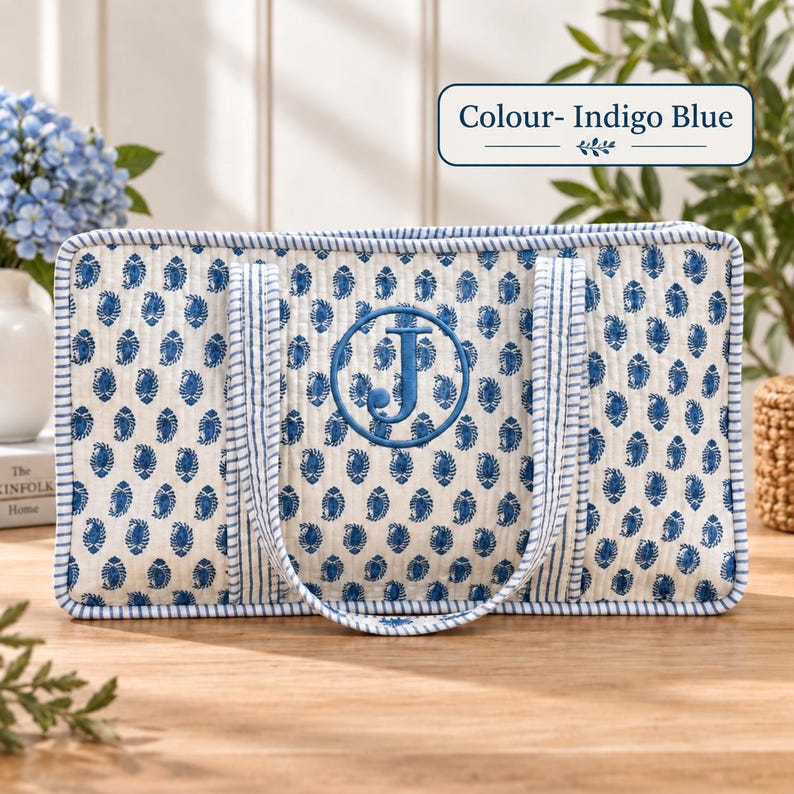 May include: A rectangular quilted tote bag in off-white featuring a repeating indigo blue paisley pattern. The bag has blue and white striped trim and handles, with a large embroidered indigo blue letter J inside a circle. Text reads Colour- Indigo Blue.