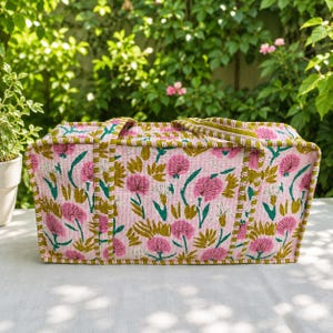 May include: A rectangular quilted fabric bag featuring a light pink base with a repeating pattern of pink flowers and green leaves. The bag includes two matching fabric handles and is finished with a green and white striped trim along the edges.