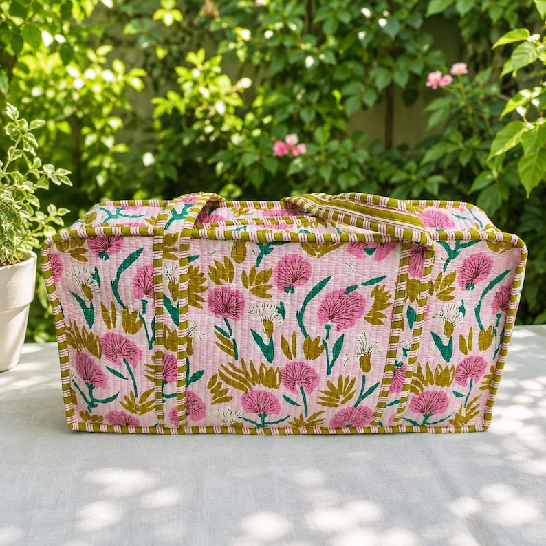 May include: A rectangular quilted fabric bag featuring a light pink base with a repeating pattern of pink flowers and green leaves. The bag includes two matching fabric handles and is finished with a green and white striped trim along the edges.