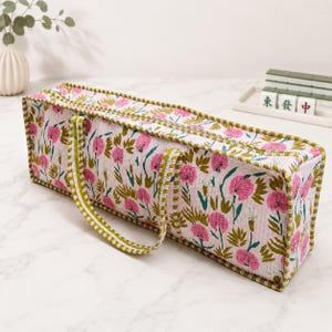 May include: A rectangular quilted fabric bag featuring a floral pattern with pink flowers and green leaves on a white background. The bag has a zippered top closure and two green and white striped fabric handles attached to the sides.