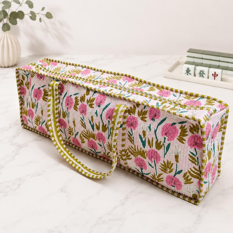 May include: A rectangular quilted fabric bag featuring a floral pattern with pink flowers and green leaves on a white background. The bag has a zippered top closure and two green and white striped fabric handles attached to the sides.