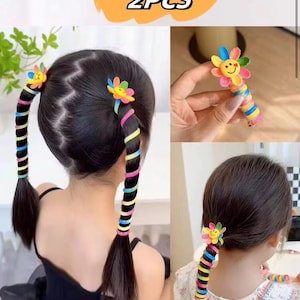 May include: Two hair accessories featuring a yellow smiley face flower topper with multi-colored petals. Each accessory has a spiral wrap design in alternating blue, pink, yellow, and green colors that coils around a ponytail. The text 2PCS is visible.