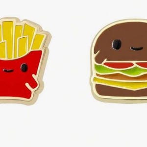 May include: Two enamel pins featuring food characters with simple faces. One pin is a red container of yellow french fries, and the other is a brown hamburger with layers of red tomato, green lettuce, and yellow cheese, both with gold-toned metal edges.