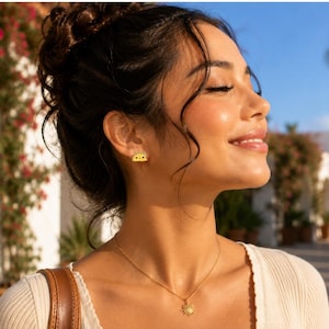 May include: A small yellow taco-shaped stud earring with a smiling face and a gold-toned necklace featuring a sunburst pendant. The earring is worn on the earlobe, and the necklace rests on the collarbone, showcasing a delicate metal chain design.