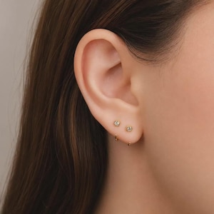 Op de afbeelding: Twee kleine gouden oorknopjes met heldere ronde stenen in een zetting, geplaatst boven een dunne gouden oorring. De sieraden worden in de oorlel gedragen en tonen een delicaat en eenvoudig ontwerp met gepolijst metaal en fonkelende stenen.