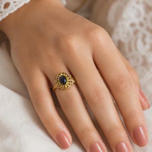 May include: A gold-toned ring featuring an oval-shaped dark blue stone at the centre. The setting is decorated with small metallic spheres and leaf-like motifs on the sides of the band, creating a detailed frame around the central gemstone.