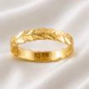 May include: A gold ring featuring a continuous band design with a raised, textured pattern resembling a series of overlapping leaves or laurel branches. The metal has a polished, reflective finish that catches the light, creating highlights along the curved surface of the band.