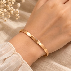 May include: A gold-toned metal cuff bracelet featuring a hammered texture across its entire outer surface. The bracelet has a slim, uniform width and a polished finish that reflects light, creating a subtle pattern of indentations along the band.