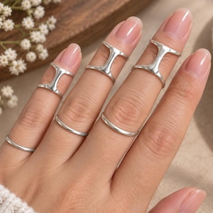 May include: A set of six silver-toned rings worn on the fingers. Three rings feature an open, H-shaped design positioned on the upper finger joints, while three plain, thin, smooth bands are worn on the lower finger joints of the same hand.