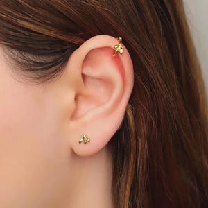 Peut inclure: Une boucle d'oreille à tige en forme d'abeille dorée placée sur le lobe de l'oreille et une boucle d'oreille créole assortie en forme d'abeille portée en piercing au cartilage. Les deux bijoux présentent un motif d'abeille détaillé.