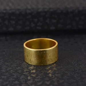 May include: A wide, circular gold-toned ring featuring a textured, matte surface with subtle variations in the finish. The band has a consistent width throughout its circumference, presenting a plain, unadorned design with a metallic, golden appearance.