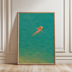 May include: A framed vertical wall art print featuring a blonde doll wearing a pink swimsuit, positioned in the center of a solid teal background. The doll is depicted in a swimming pose with arms outstretched, surrounded by a vast expanse of deep teal.