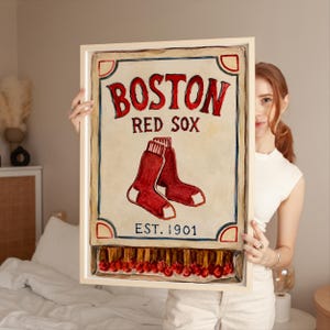 May include: A framed rectangular wall art piece featuring a vintage-style matchbox design with the text BOSTON RED SOX and EST. 1901. The illustration displays two red socks in the centre and a row of match heads at the bottom in a light frame.
