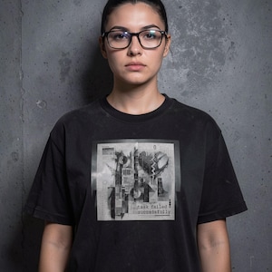 Puede incluir: Una camiseta negra de manga corta con un estampado gráfico cuadrado en el pecho. El diseño incluye formas geométricas abstractas, un icono de corazón roto y el texto "task failed successfully" en una fuente pequeña y sin remates.