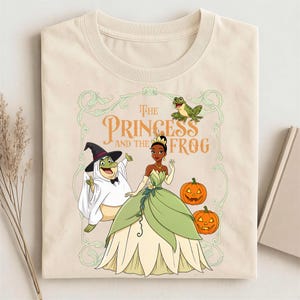 Puede incluir: Una camiseta de color crema con un estampado gráfico de Tiana con un vestido verde, una rana con sombrero de bruja y una rana pequeña. El texto dice The Princess and the Frog, rodeado por un marco ornamentado verde claro y dos calabazas naranjas.