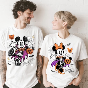 Puede incluir: Dos camisetas blancas con gráficos de Halloween. La camiseta de la izquierda muestra a Mickey Mouse con un disfraz de esqueleto, capa morada, calabazas y una telaraña, con el texto Mickey Mouse. La de la derecha muestra a Minnie Mouse con un disfraz de esqueleto, capa morada, calabazas y el texto Spooky Vibes.