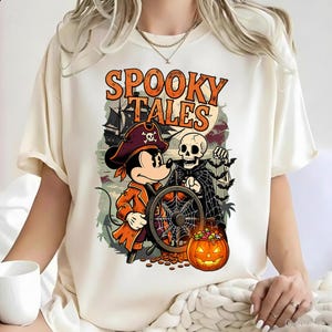 Puede incluir: Una camiseta color crema con un estampado gráfico que incluye el texto "SPOOKY TALES" en naranja. El diseño muestra a un ratón de dibujos animados vestido de pirata sosteniendo un timón, un esqueleto con capa negra y una calabaza.